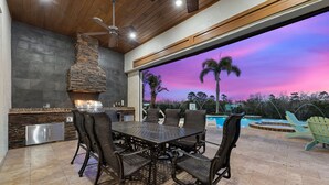 Cocina exterior con chimenea de piedra, mesa de comedor con sillas y vista a una piscina al atardecer.