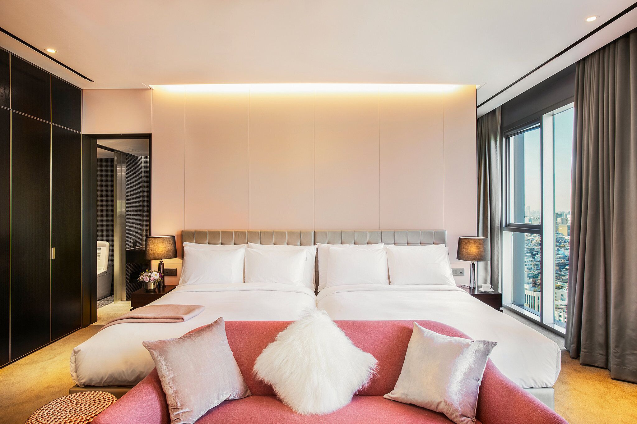 A modern hotel room with a large bed, a red sofa, and a view of the city through the window.