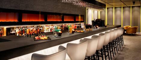 Una zona de bar con una barra alargada, varias botellas de licores, vasos y una variedad de alimentos. Hay varios taburetes y un escenario al fondo.