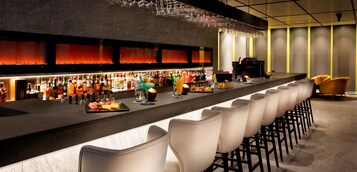 Una zona de bar con una barra alargada, varias botellas de licores, vasos y una variedad de alimentos. Hay varios taburetes y un escenario al fondo.