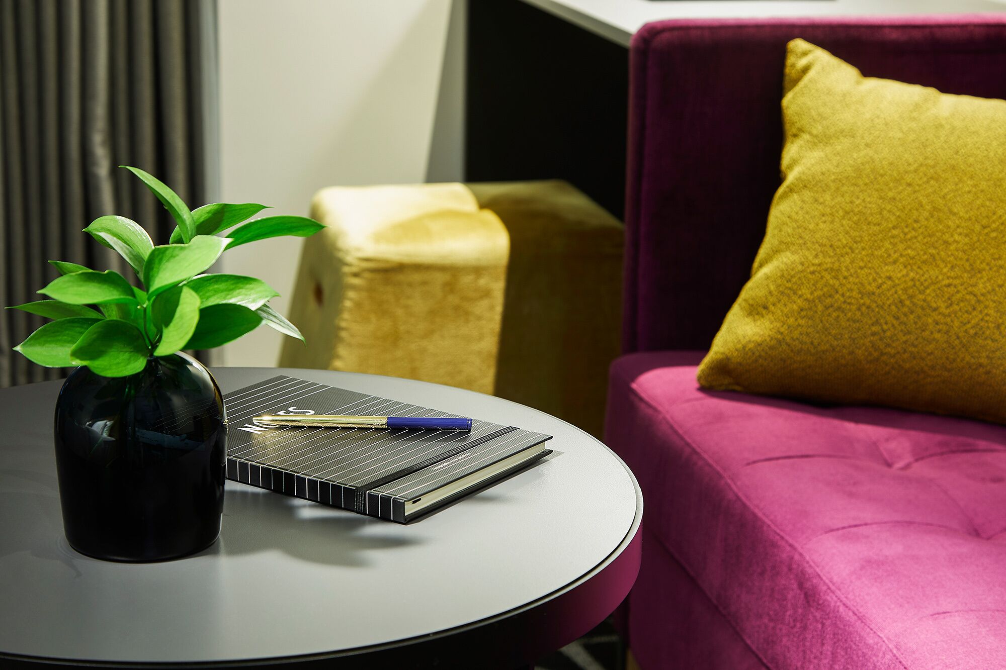 Una habitación moderna con una mesa redonda, una planta en maceta, un cuaderno y un cojín amarillo sobre un sofá morado.