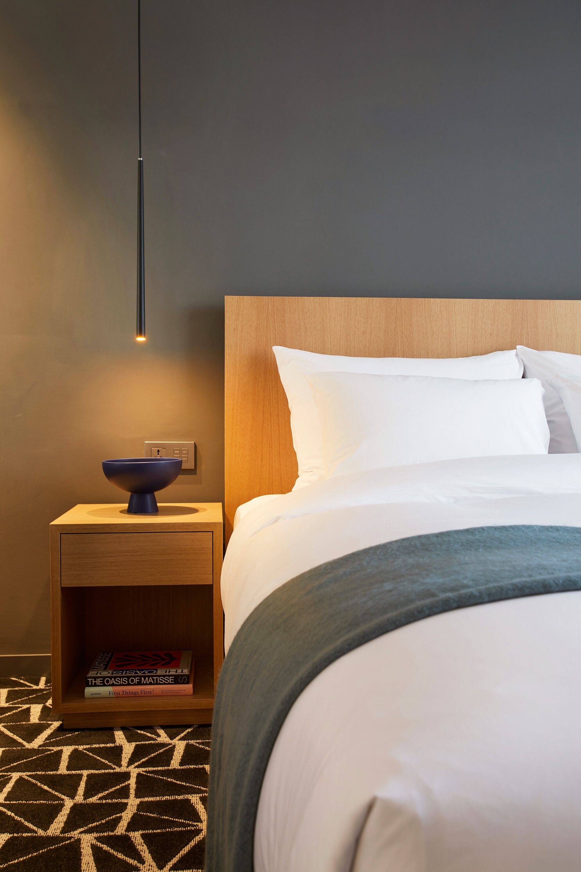 Una cama bien hecha con ropa blanca, un cabecero de madera y una mesita de noche con un cuenco azul y libros.