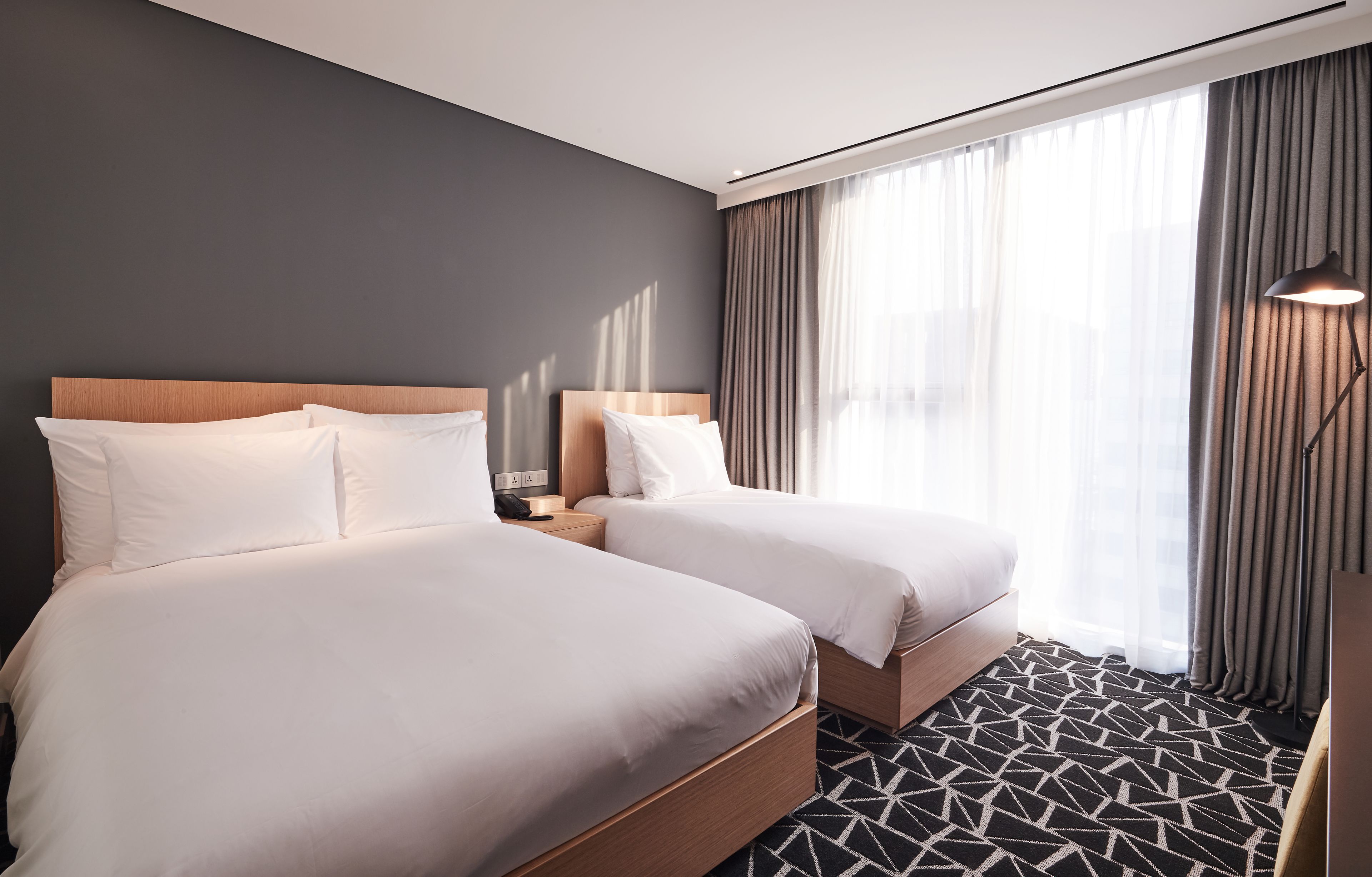 A hotel room with two beds, a modern design, a geometric patterned carpet, and a large window with curtains.