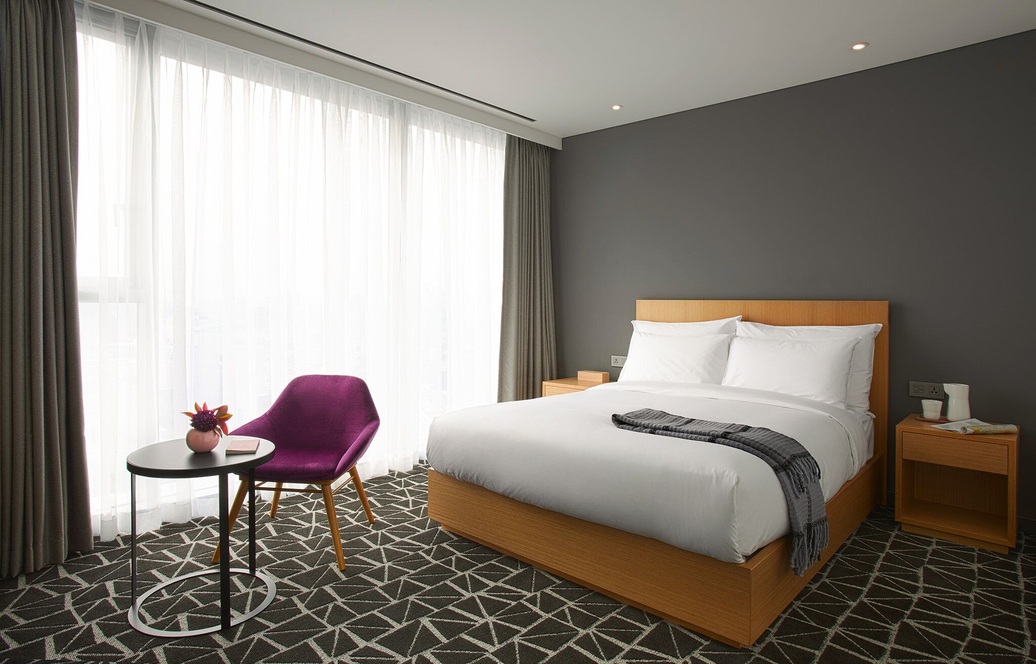 A hotel room with a bed, a small table, a chair, and a nightstand.