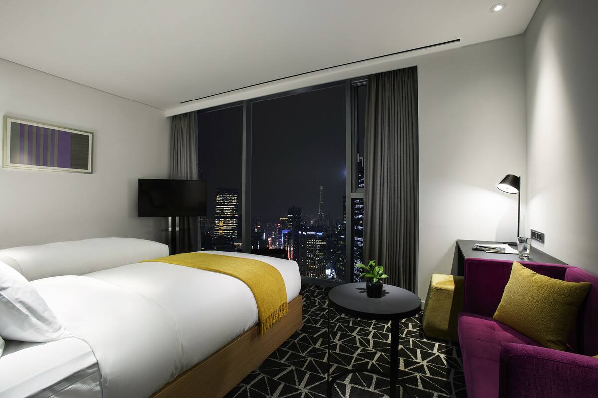 Une chambre d’hôtel avec un grand lit, un petit canapé, une table ronde et une vue sur le paysage urbain illuminé la nuit.