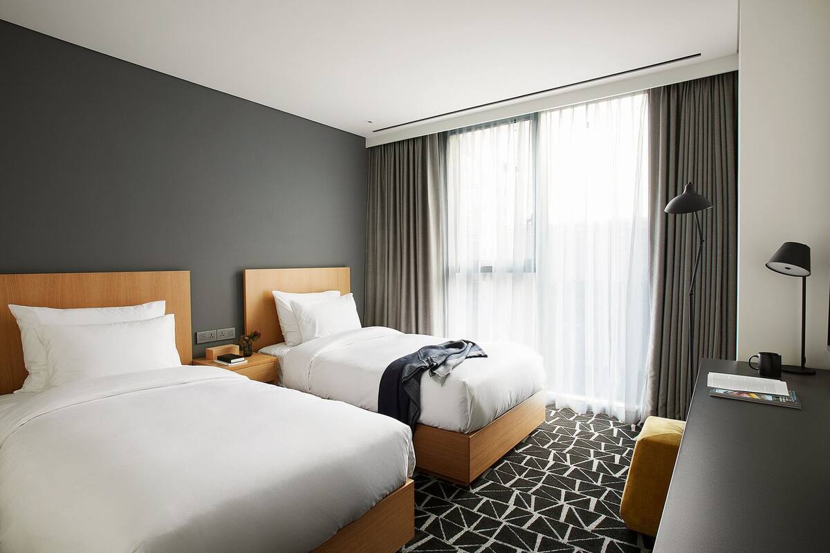Une chambre d’hôtel avec deux lits, un bureau, une chaise et une grande fenêtre avec des rideaux.