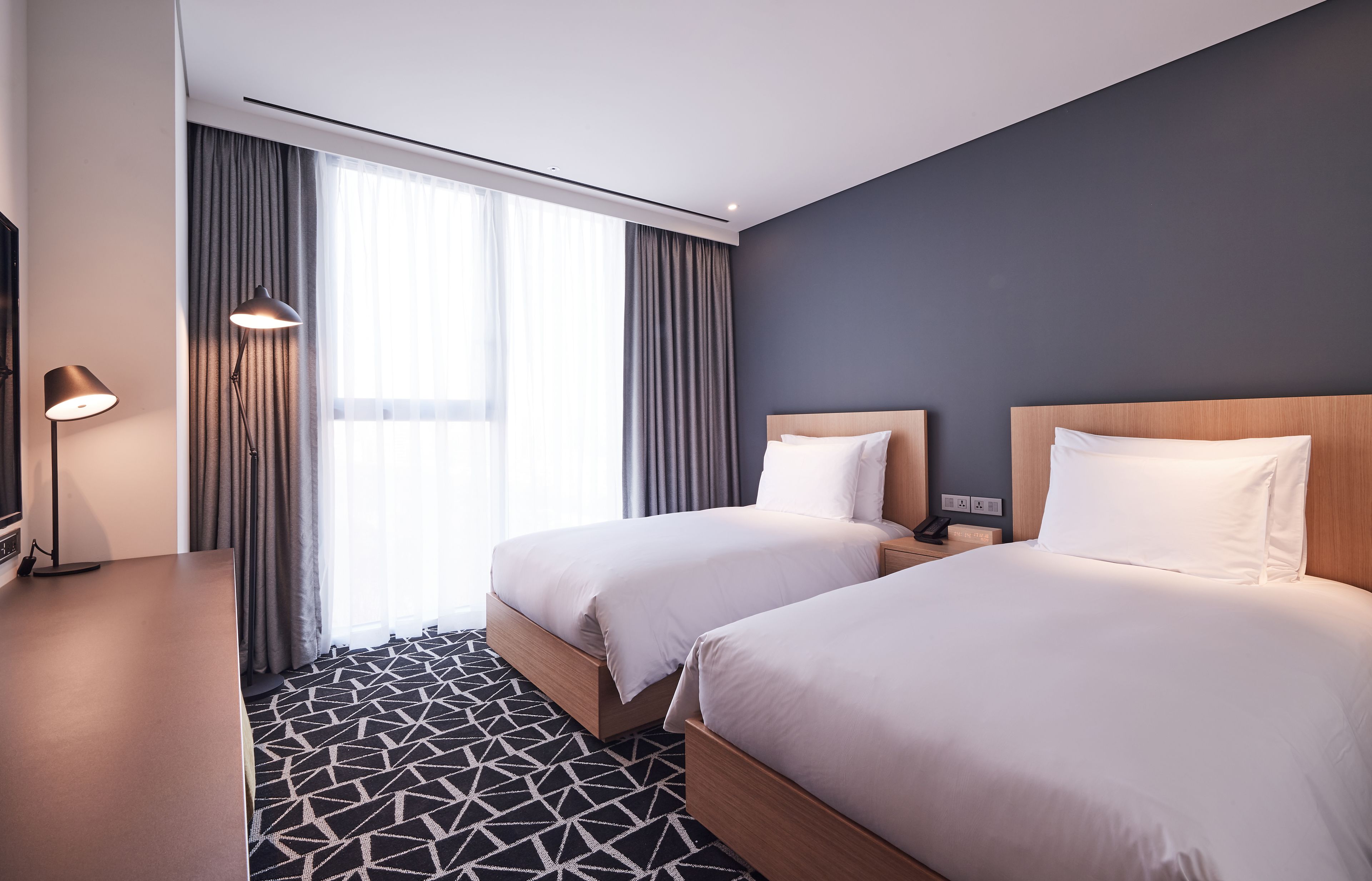 Habitación de hotel con dos camas, un escritorio, una lámpara y un ventanal grande con cortinas.