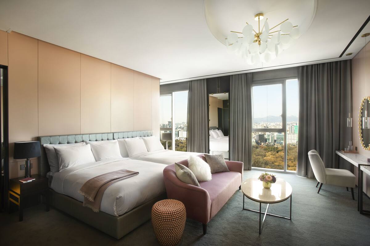 Une chambre d’hôtel moderne avec un grand lit, un canapé, une petite table avec un vase, et une vue sur la ville grâce à de grandes fenêtres.