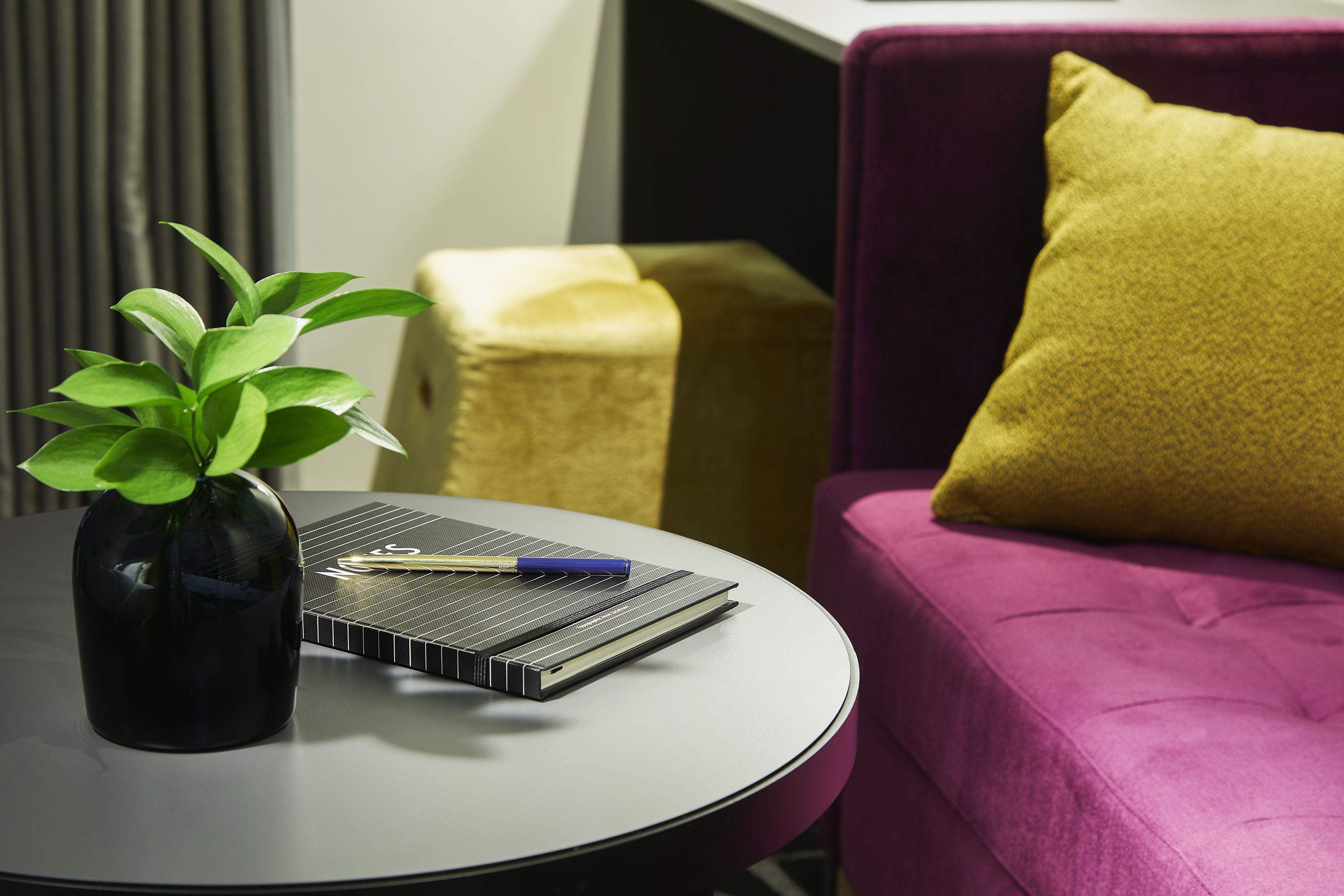A round table with a black vase containing a green plant, a notebook, and a pen. A purple sofa with a yellow pillow is in the background.