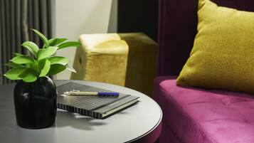 A round table with a black vase containing a green plant, a notebook, and a pen. A purple sofa with a yellow pillow is in the background.