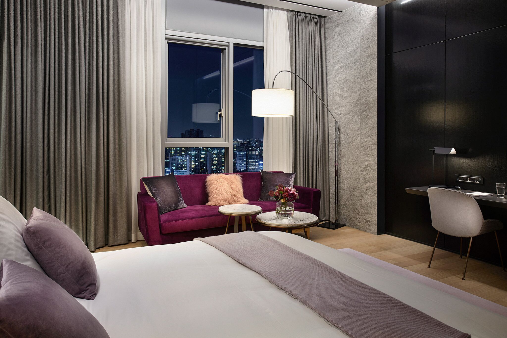 A modern hotel room with a large bed, a purple sofa, a small round table with a vase of flowers, a desk with a chair, and a city view through the window.
