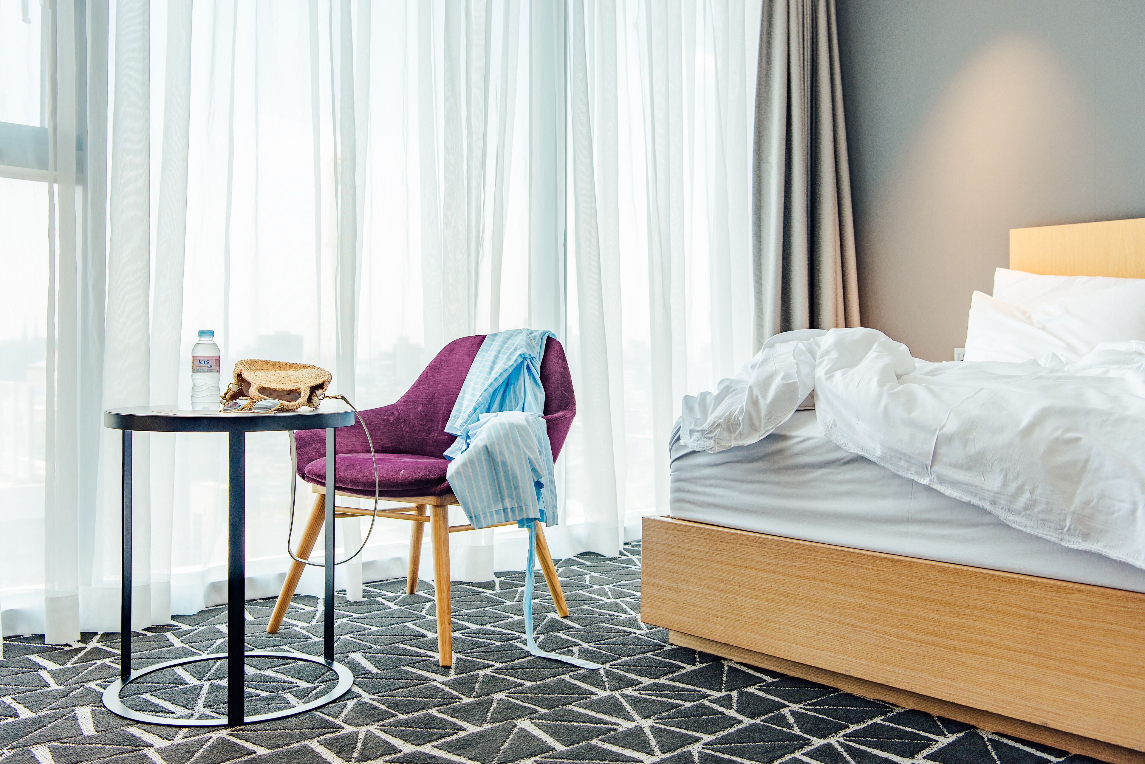 A hotel room with a bed, a chair, a small table, and a window with sheer curtains.
