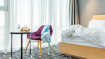 A hotel room with a bed, a chair, a small table, and a window with sheer curtains.