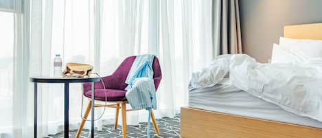 Habitación de hotel con una cama, una silla, una mesita y una ventana con cortinas traslúcidas.