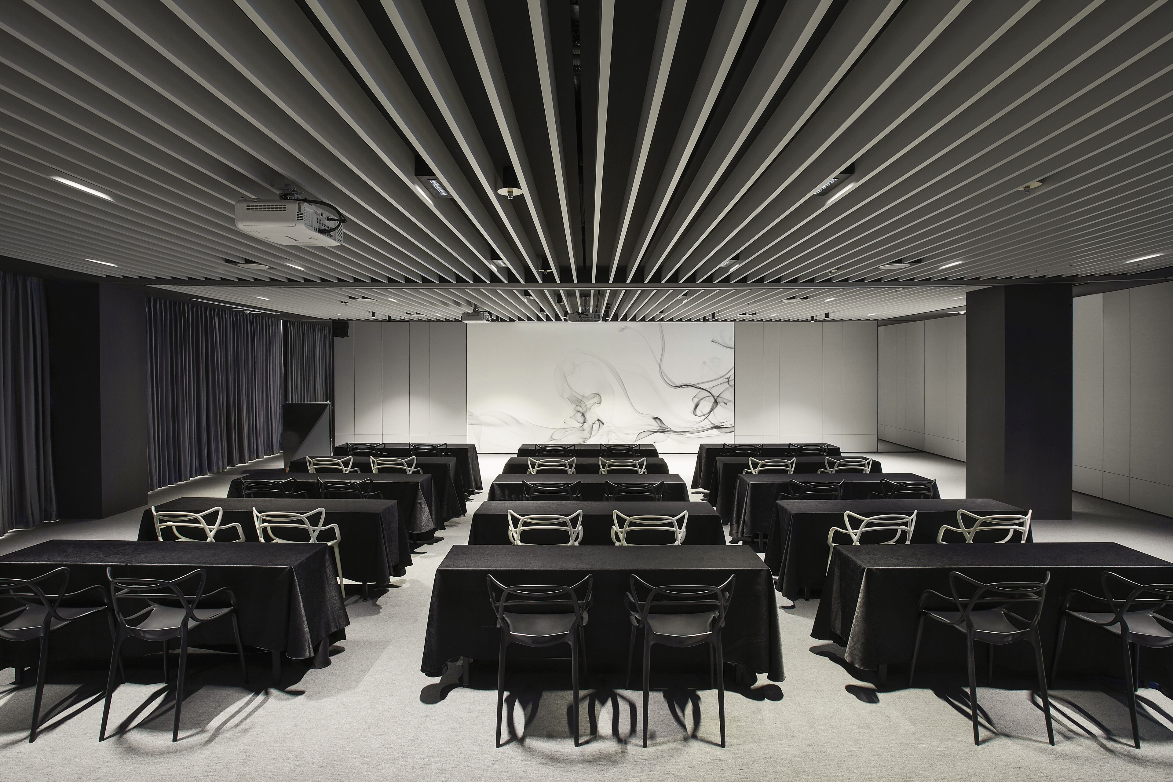 Un salón de conferencias con filas de mesas y sillas, un proyector y una gran obra de arte abstracto en la pared.