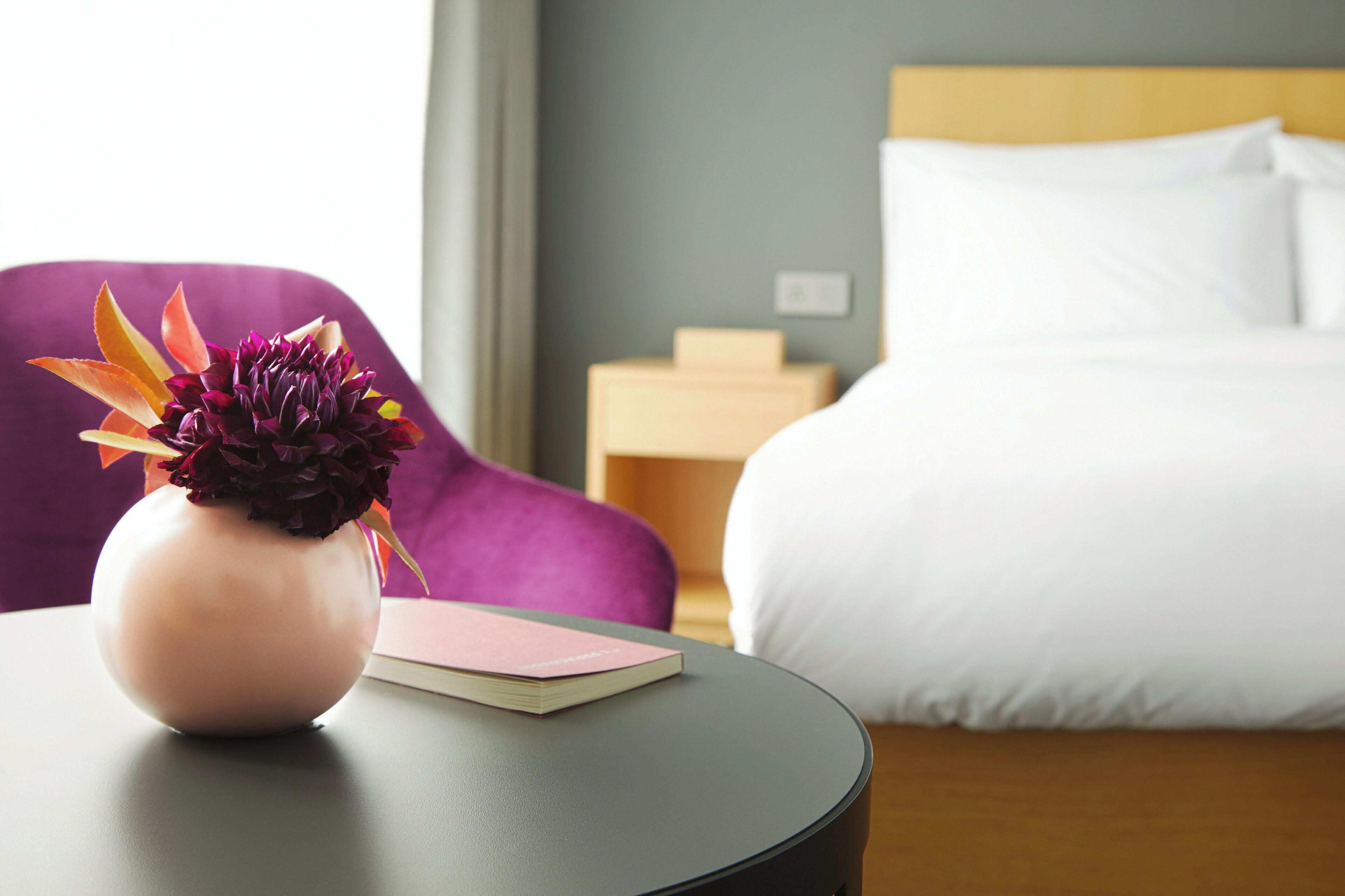 A hotel room with a bed, a bedside table with a vase of flowers, a purple chair, and a small wooden nightstand.