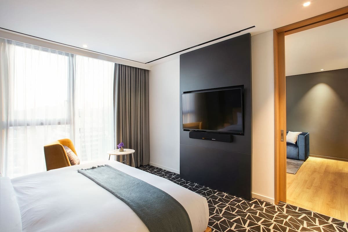 A modern hotel room with a large bed, a television mounted on the wall, a small table with a vase, and a view of the city through large windows.