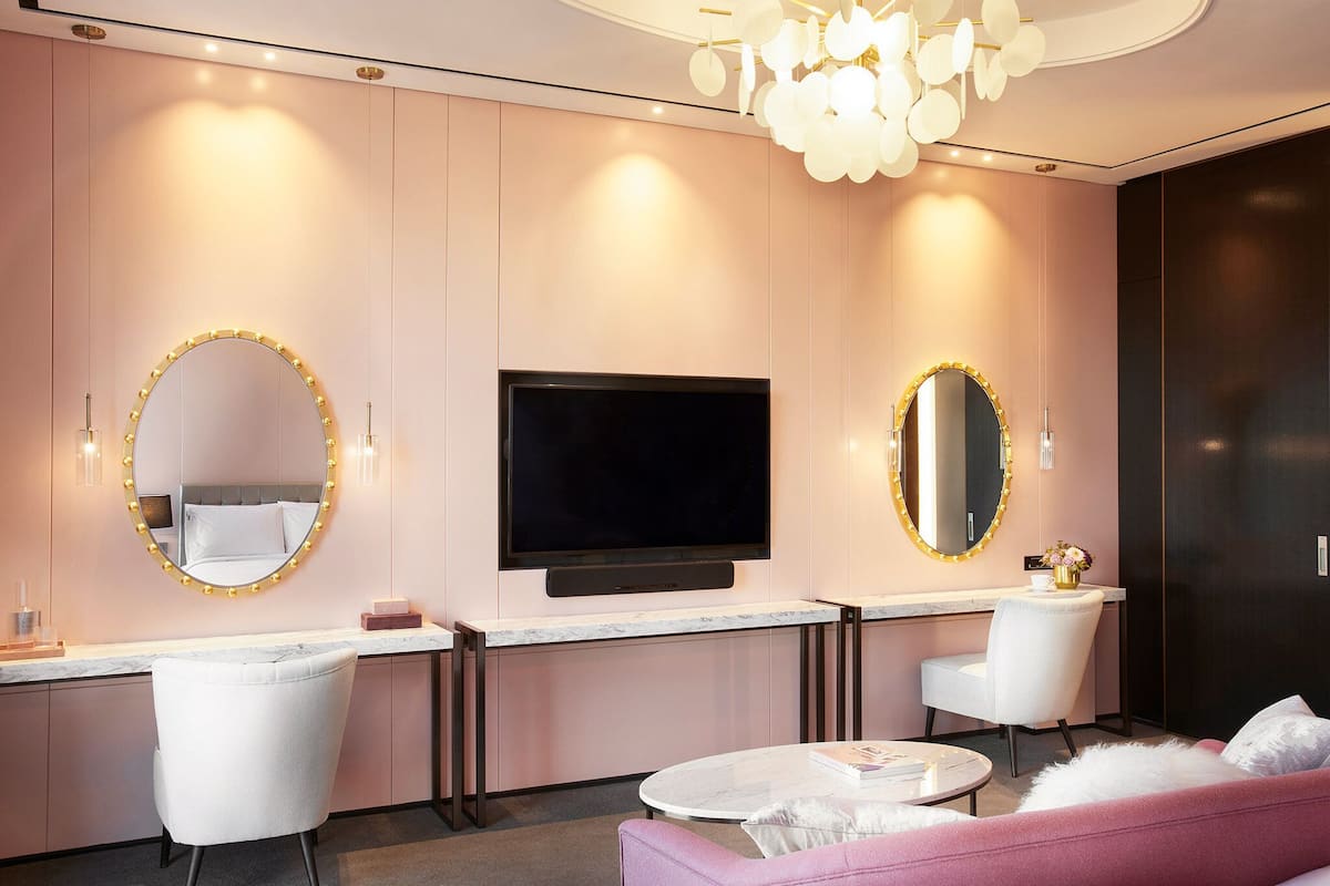 A modern hotel room with a large flat-screen TV mounted on the wall, a vanity with a mirror, and a pink sofa.