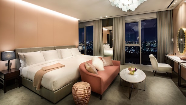 Een moderne hotelkamer met een groot bed, een roze bank, een ronde tafel en uitzicht op de stad door grote ramen.