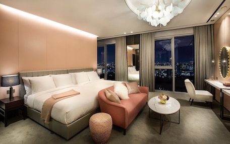 A modern hotel room with a large bed, a pink sofa, a round table, and a city view through large windows.