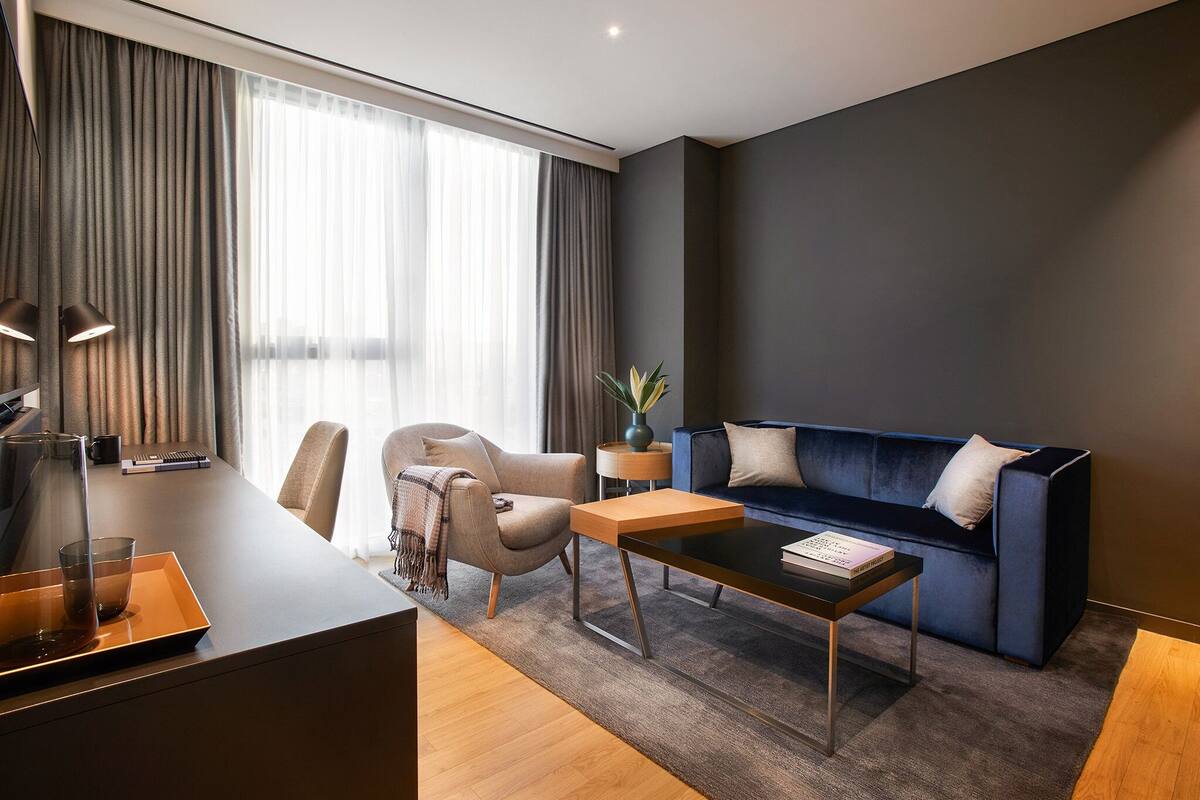 Une chambre d’hôtel moderne avec un canapé bleu foncé, une table basse en bois, un fauteuil beige et un bureau avec une lampe.