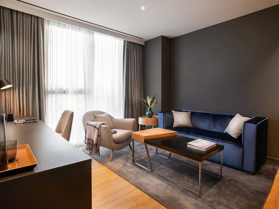 Una habitación de hotel moderna con un sofá azul oscuro, una mesa de centro de madera, un sillón color beige y un escritorio con lámpara.