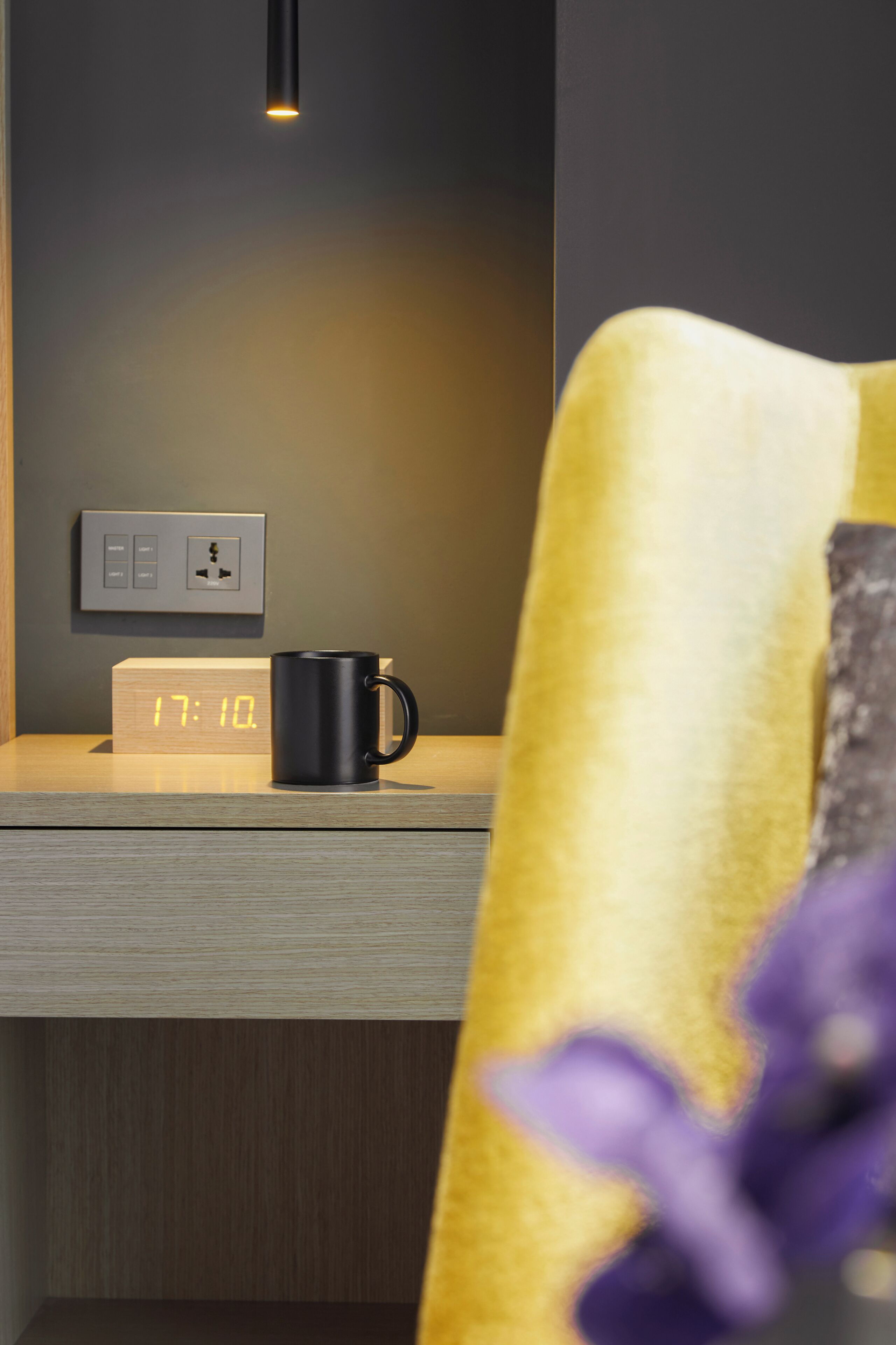 A wooden bedside table with a digital clock displaying 17:10, a black mug, and a wall socket.