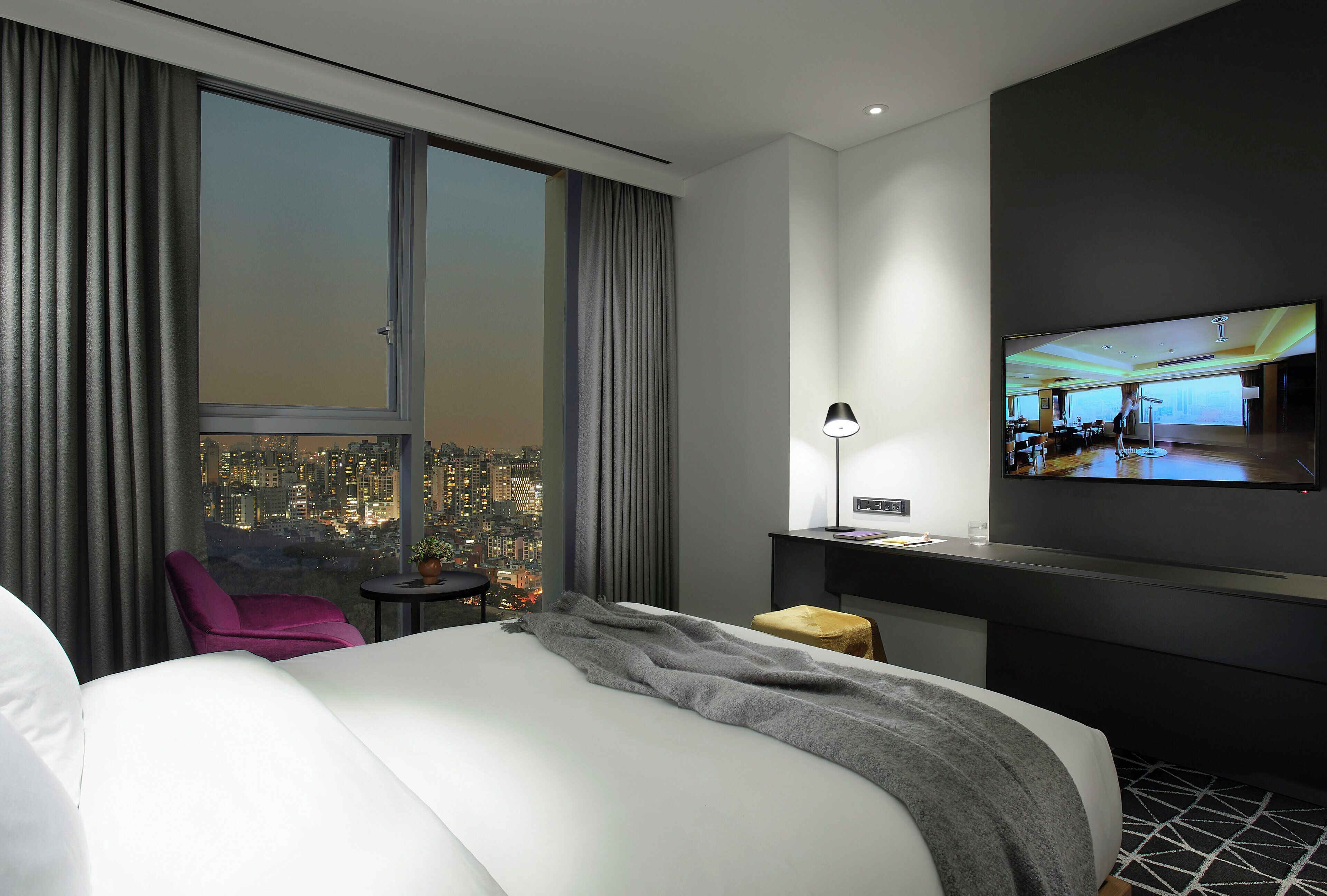 A modern hotel room with a large bed, a desk, a chair, and a TV mounted on the wall.