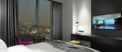 A modern hotel room with a large bed, a desk, a chair, and a TV mounted on the wall.