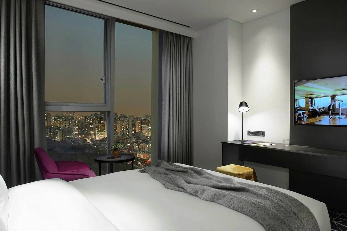 Une chambre d’hôtel avec un grand lit, un bureau avec une lampe, un téléviseur fixé au mur et une vue sur le paysage urbain.