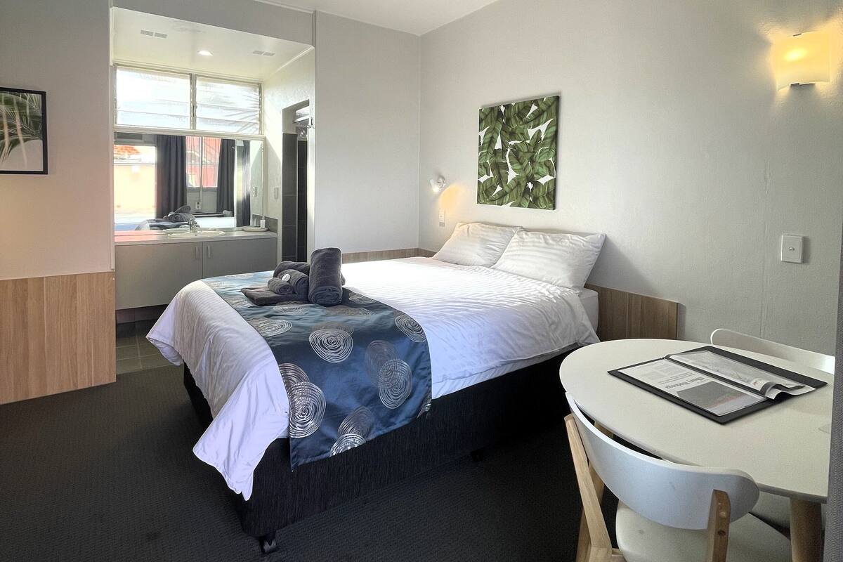 A hotel room with a bed, a desk, and a chair. There is a window and a door visible.