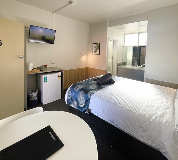 Habitación de hotel con cama, escritorio con computadora, refrigerador pequeño, televisor montado en la pared y baño visible a través de una puerta abierta.