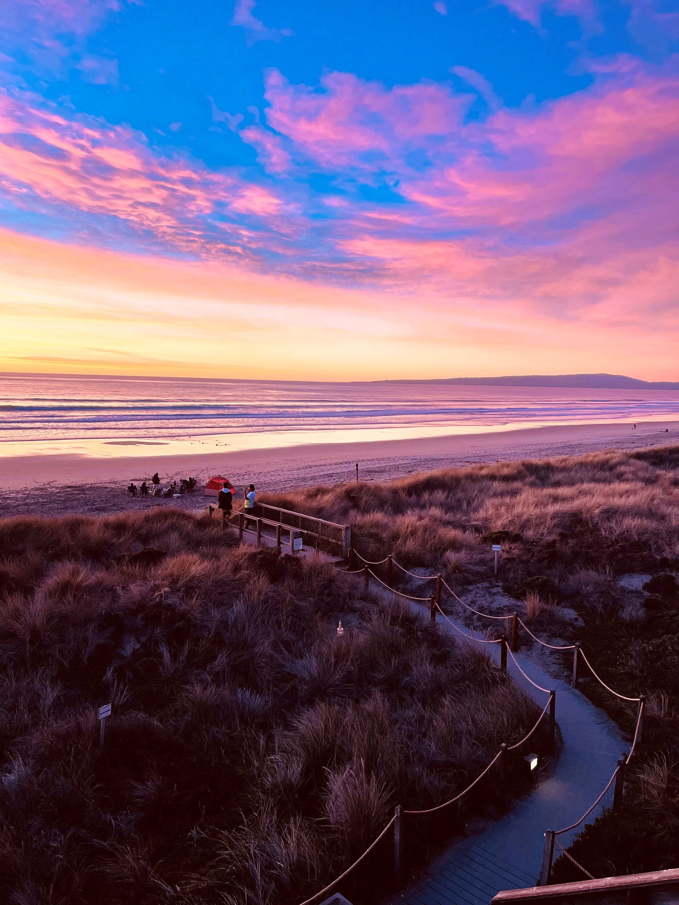A coastal landscape with a wooden boardwalk, grassy dunes, and a distant shoreline.