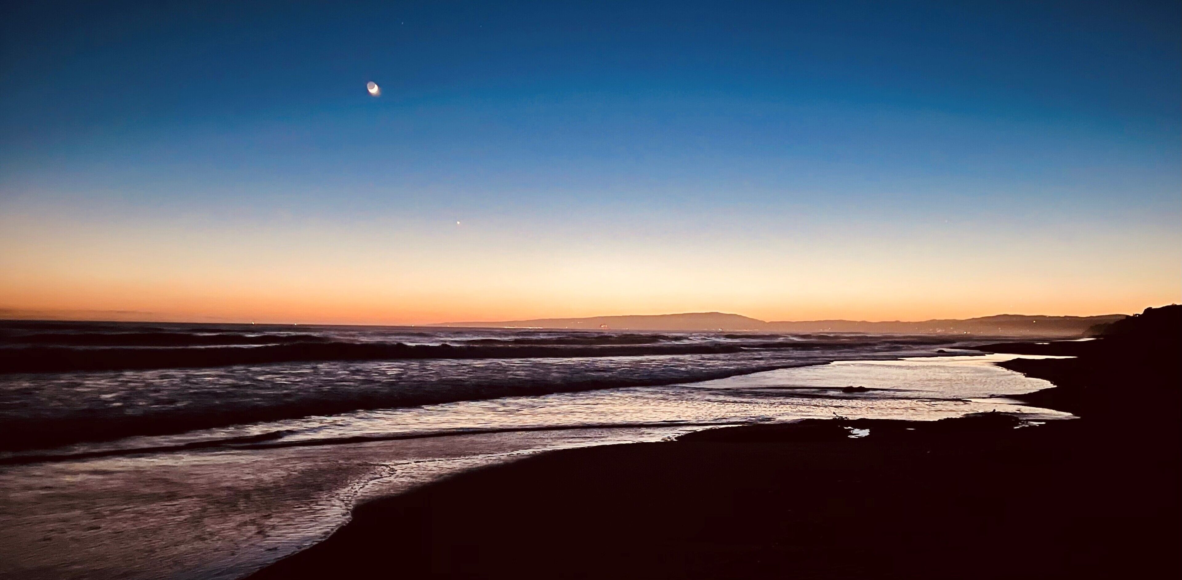 Une plage au coucher du soleil, avec des vagues, une lune en croissant et des montagnes à l’horizon.