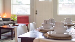 A dining area with a table set for four, white tableware, and a view of the outdoors through large windows.