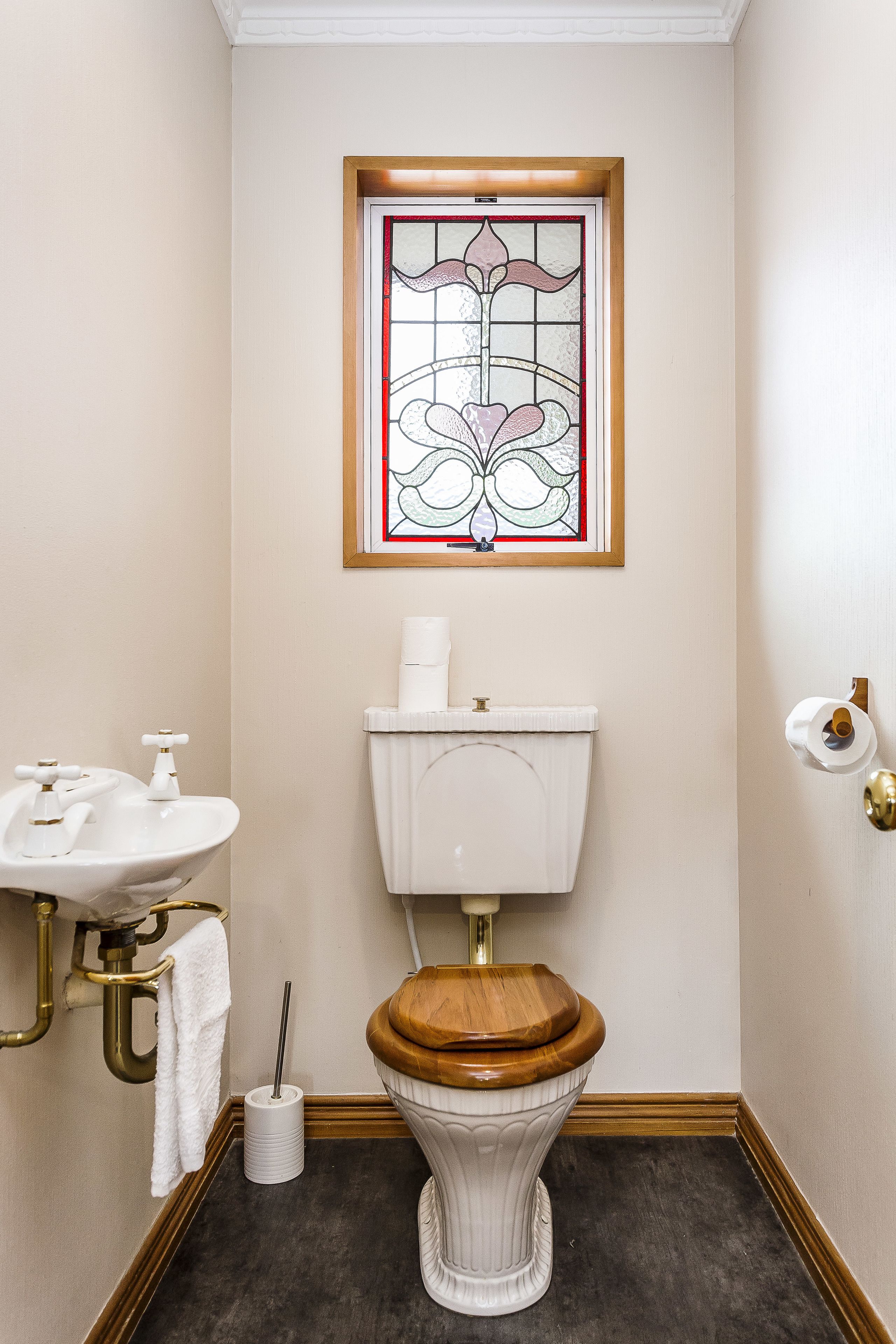 A bathroom with a white toilet, a wooden toilet seat, a white sink with a gold faucet, a roll of toilet paper, and a stained glass window above the toilet.