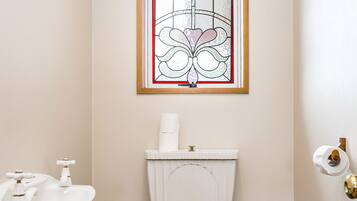 A bathroom with a white toilet, a wooden toilet seat, a white sink with a gold faucet, a roll of toilet paper, and a stained glass window above the toilet.