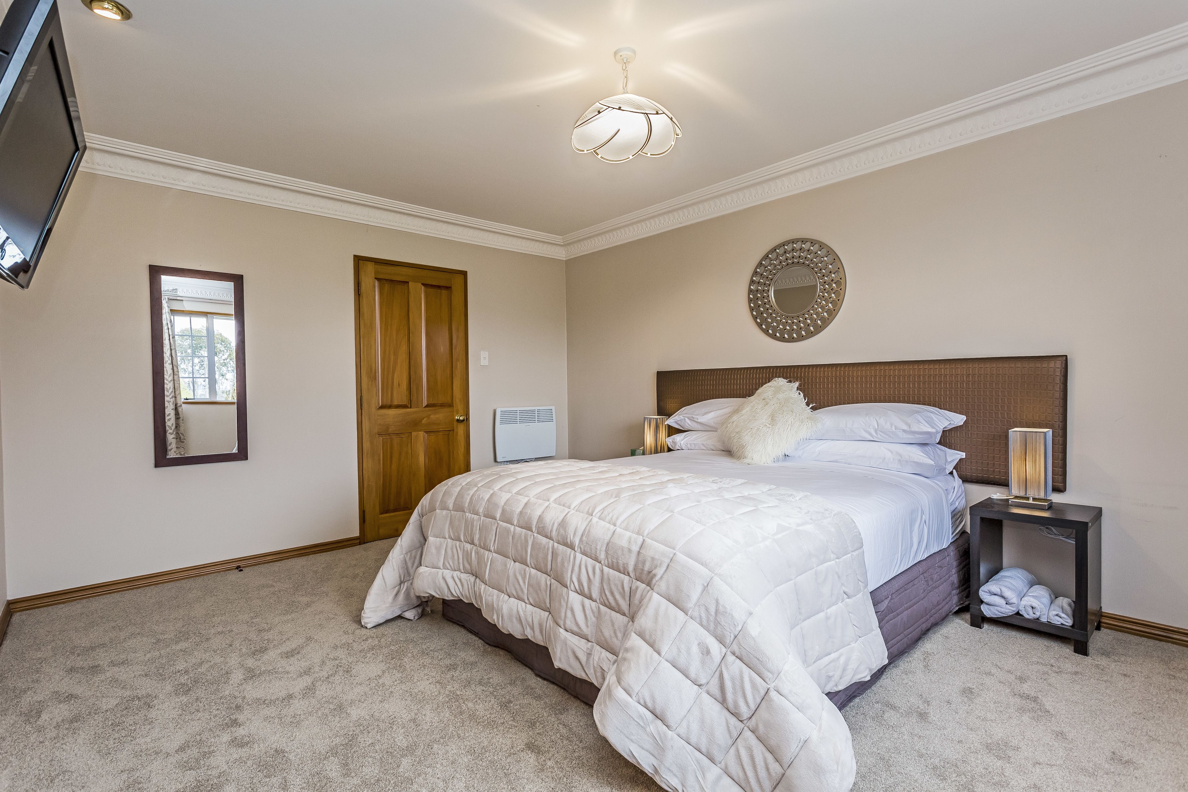 A bedroom with a large bed, a bedside table with a lamp, a mirror on the wall, and a door.