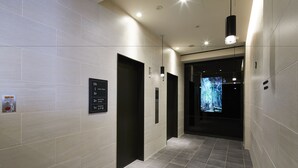 Un couloir moderne aux murs et au sol carrelés, doté de portes d’ascenseur et d’un écran numérique.