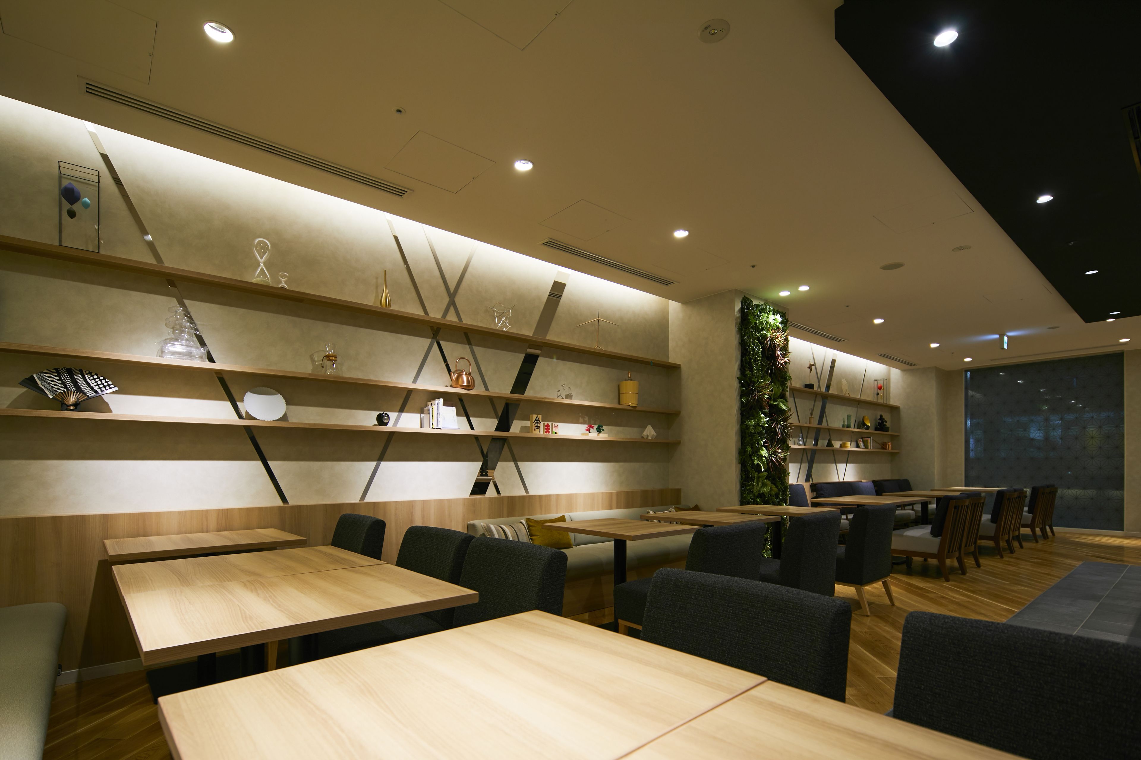 A modern restaurant with wooden tables and chairs, shelves with decorative items, and a large window with a cityscape view.
