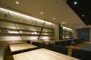 A modern restaurant with wooden tables and chairs, shelves with decorative items, and a large window with a cityscape view.