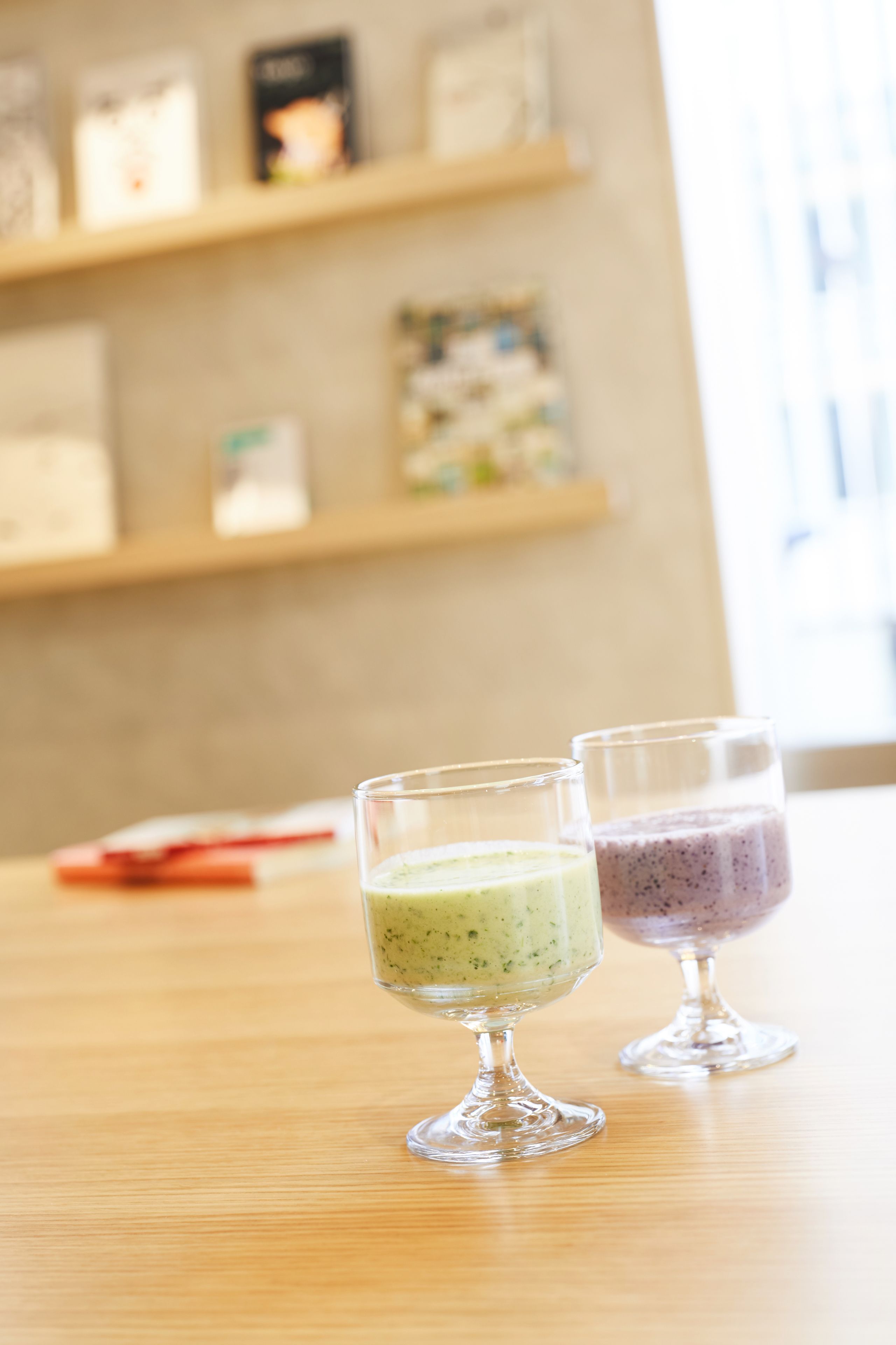 Dois copos de smoothies verdes e roxos sobre uma mesa de madeira, com um fundo desfocado de prateleiras.