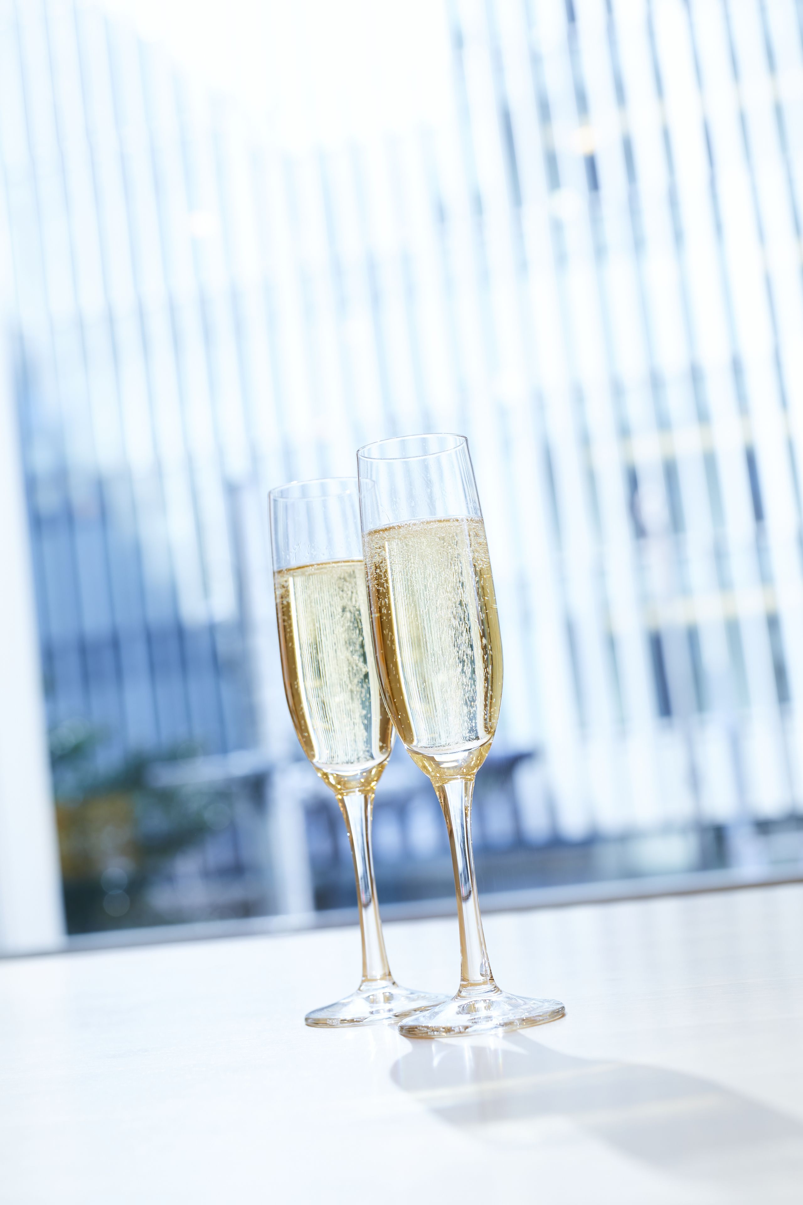 Two champagne flutes on a reflective surface with a blurred background of vertical lines, possibly a window or wall.