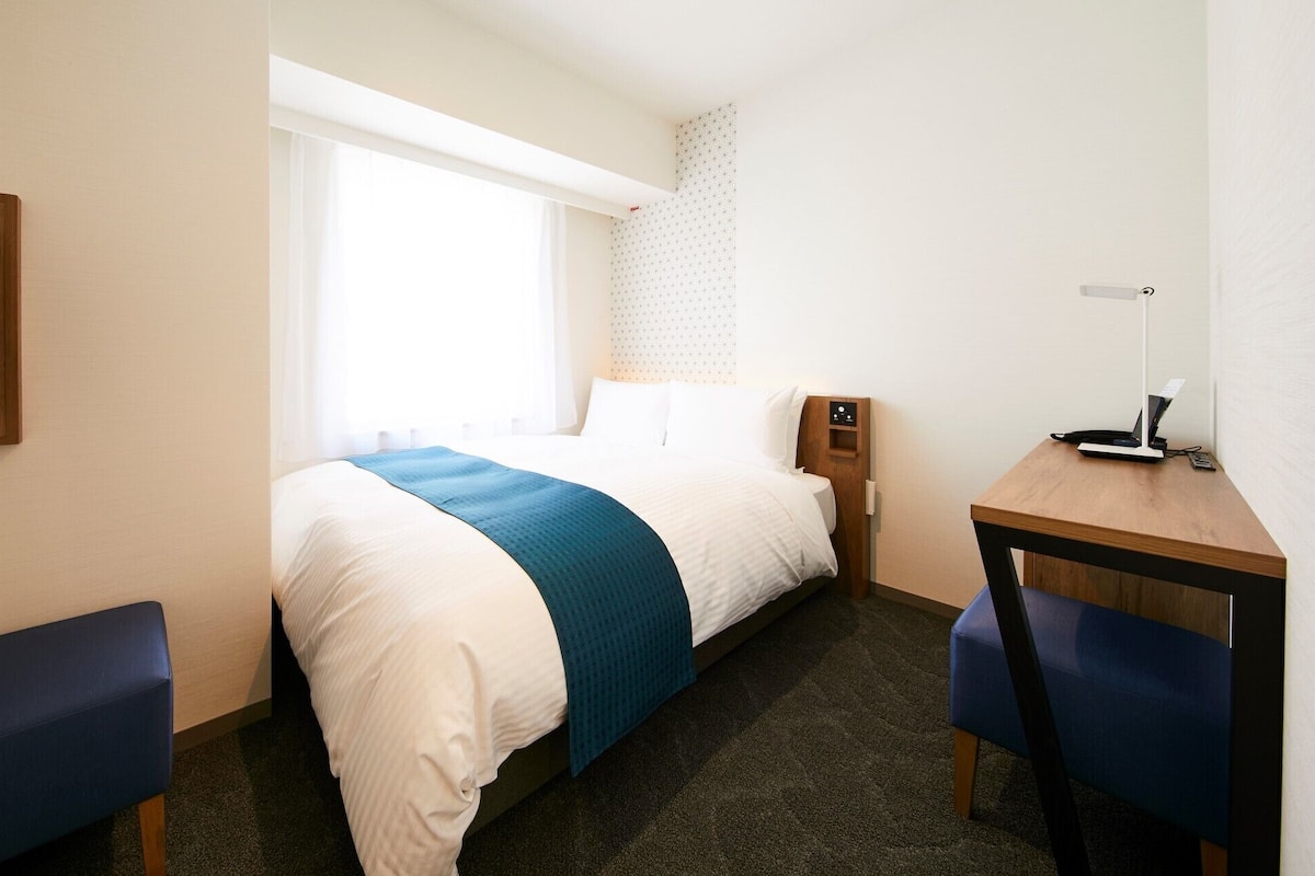 Hotellihuone, jossa on sänky, työpöytä kannettavalla tietokoneella, televisio ja ikkuna, jossa on verhot.