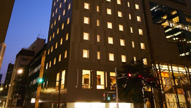 Un grand hôtel à la façade ponctuée de nombreuses fenêtres, avec une entrée bien visible, situé à un carrefour où se trouvent des feux de signalisation.