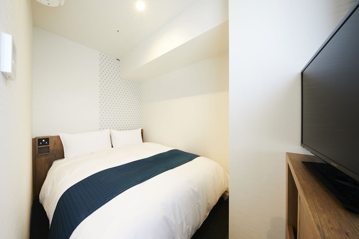 Una habitación de hotel con una cama, un televisor sobre un soporte de madera y una lámpara fijada a la pared.