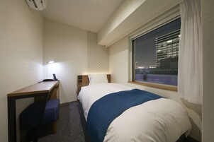 Habitación de hotel con cama, escritorio, silla, mesita de noche, lámpara y ventana con vistas a un paisaje urbano.
