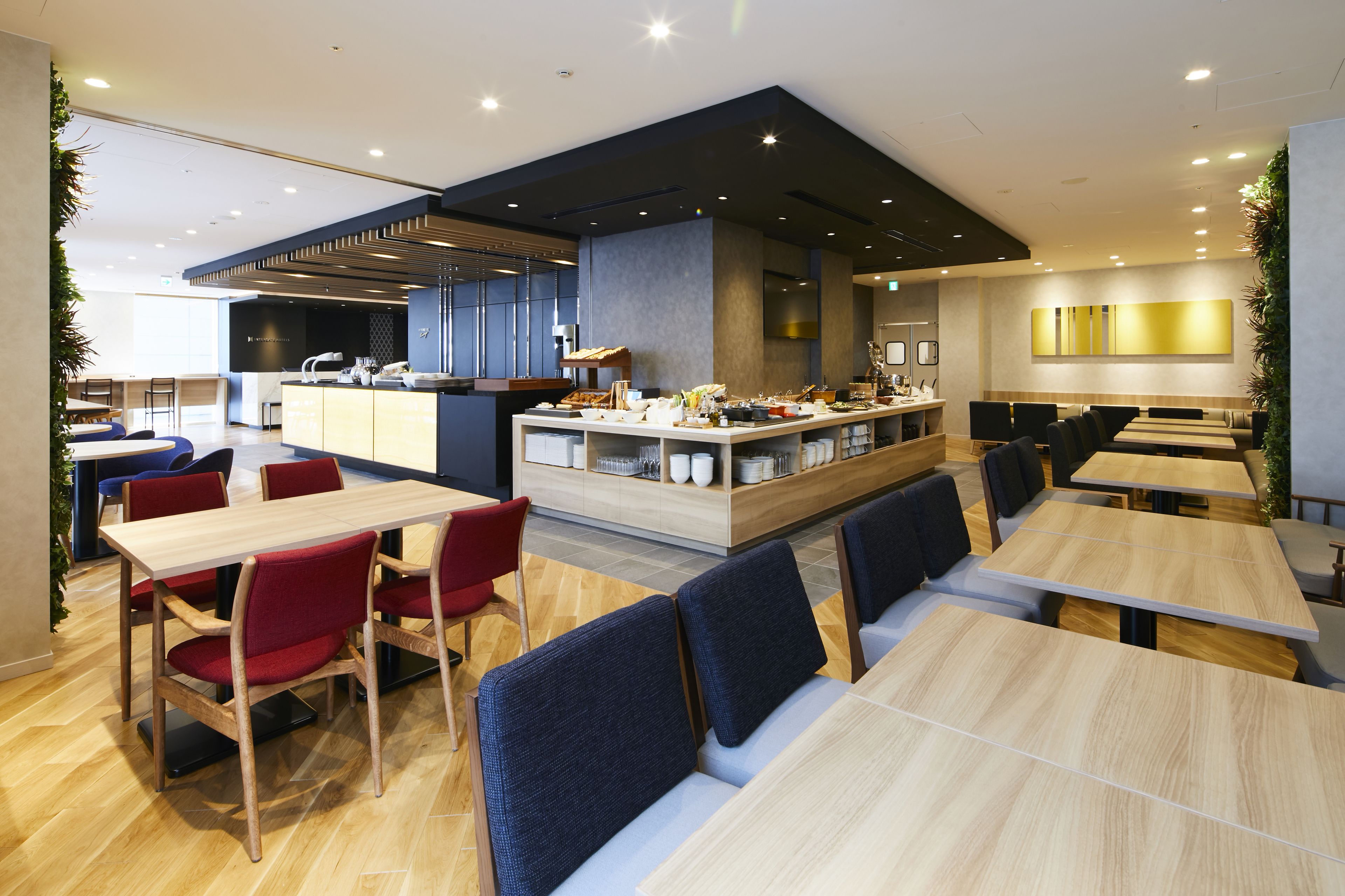 A modern restaurant with wooden tables and red chairs, a central counter with a buffet, and a bar area.