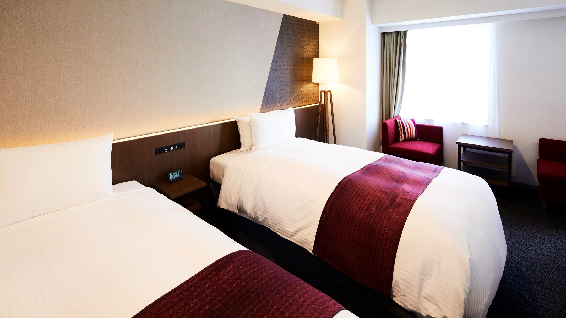 Hotelkamer met twee bedden, een nachtkastje met een klok, een lamp, een stoel en een raam met gordijnen.