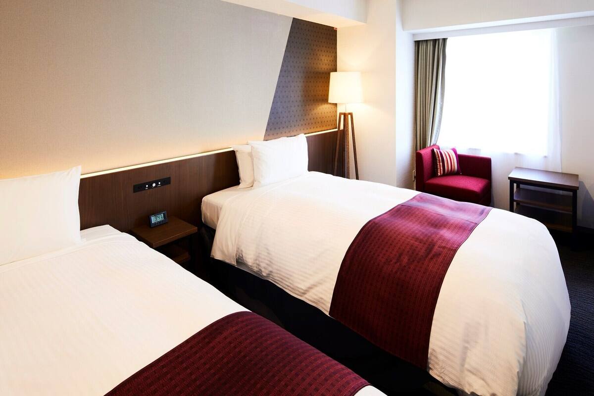 Hotellihuone, jossa on kaksi sänkyä, yöpöytä, jossa on kello, lamppu, tuoli ja ikkuna, jossa on verhot.